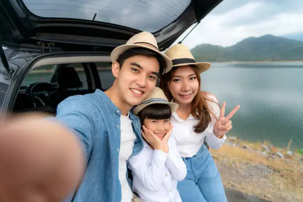 Photo of Portrait of Asian family sitting in car with father, mother and daughter selfie with lake and mountain view by smrtphone while vacation together in holiday. Happy family time.