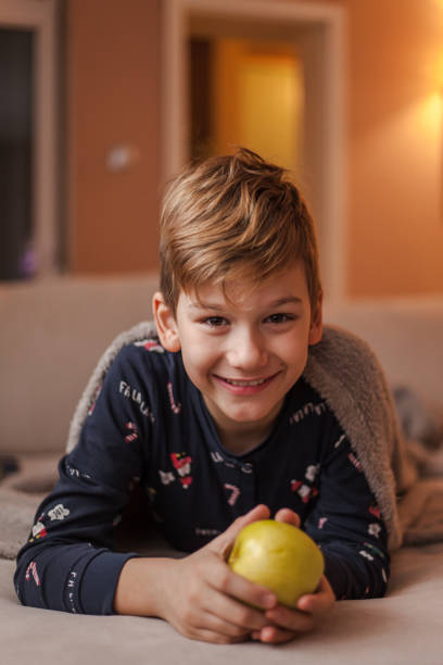 istock Cute boy holding an apple while resting on sofa in the living room. 1202910418