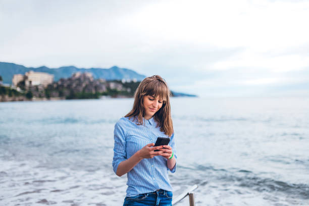 Young Caucasian beautiful woman using smartphone at seaside, with sea water behind her.