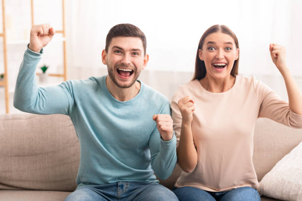 istock Joyful Couple Shouting Holding Hands Celebrating Victory Sitting On Couch 1202923758