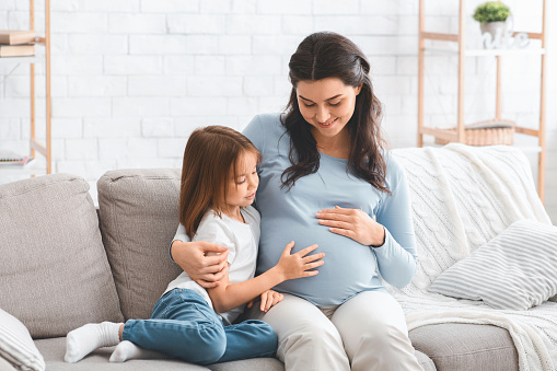 Happy pregnant mother and preschool daughter sitting on sofa at home, having conversation, copy space
