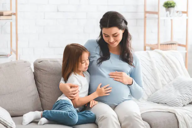 Photo of Pregnant mother and preschool daughter sitting on sofa at home