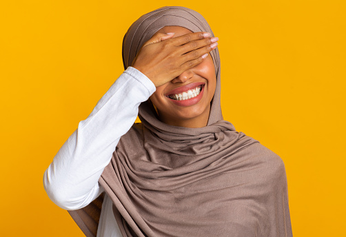 Facepalm. Cheerful Black Muslim Woman In Headscarf Covering Eyes With Hand And Laughing, Feeling Shy, Standing Over Yellow Background, Copy Space