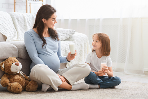 Healthy expecting young mom and little cute daughter having glass of milk, sitting on floor at living room