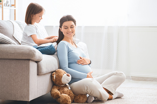 Cute little girl brushing her smiling pregnant mom hair, home interior, free space