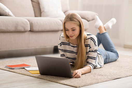 Blonde girl student studying at home, laying on floor and using laptop, empty space