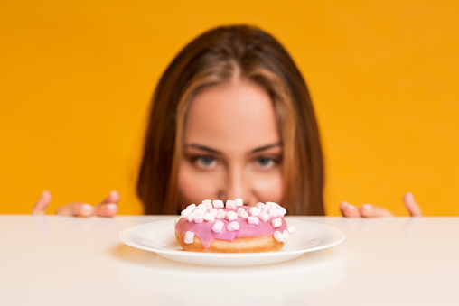 Im On Diet. Hungry woman peeking out of behind table and looking at donut with appetite, yellow studio background with empty space, selective focus