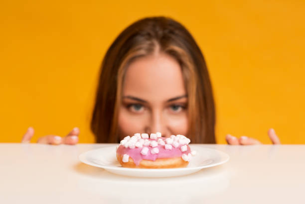 Hungry woman looking at donut with appetite, peeking out of table Im On Diet. Hungry woman peeking out of behind table and looking at donut with appetite, yellow studio background with empty space, selective focus desire stock pictures, royalty-free photos & images