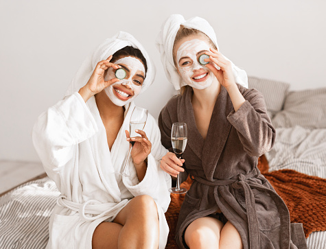 Cheerful multiracial girlfriends with face masks on wearing bathrobes drinking champagne, holding cucumber slices over eyes and smiling at camera, home interior