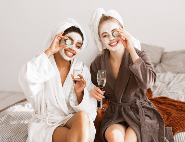 Cheerful girlfriends with face masks wearing bathrobes drinking champagne Cheerful multiracial girlfriends with face masks on wearing bathrobes drinking champagne, holding cucumber slices over eyes and smiling at camera, home interior bathrobe photos stock pictures, royalty-free photos & images