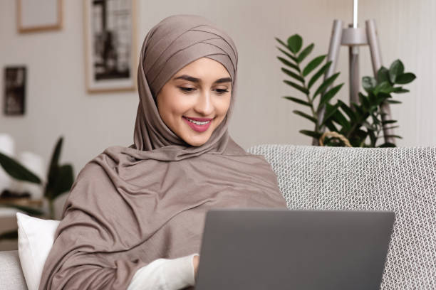 istock Girl in headscarf using laptop while sitting on sofa at home 1202956519