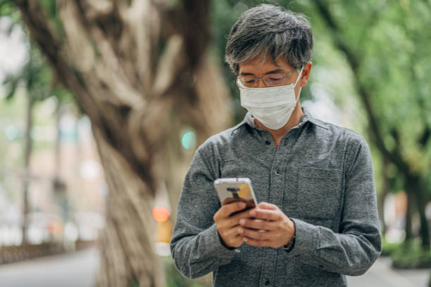 istock Man with surgical mask protecting himself from corona virus using mobile on the street 1202956536