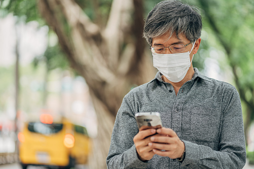 One man, mature man with surgical mask protecting himself from corona virus, using mobile phone downtown in city.