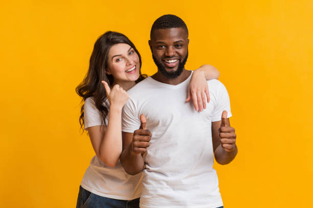 Happy millennial couple embracing and gesturing thumbs up at camera Happy millennial couple embracing and gesturing thumbs up at camera, posing together over yellow background in studio, free space black man white woman couple stock pictures, royalty-free photos & images