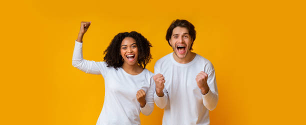 Happy interracial couple rejoicing success, celebrating victory with raised fists Big Win. Happy interracial couple rejoicing success, celebrating victory with raised fists, exclaiming with joy over yellow background, panorama clingy girlfriend stock pictures, royalty-free photos & images