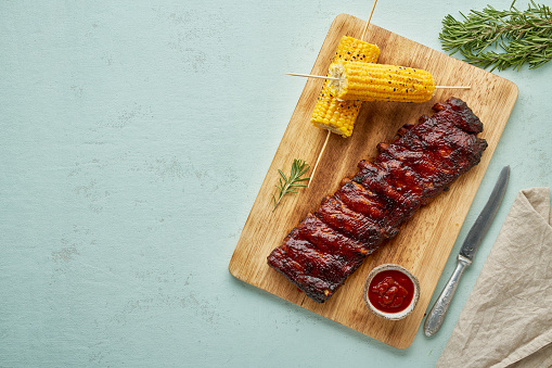 Barbecue pork ribs. Slow cooking recipe. Whole pickled roasted pork meat with red sauce and corn on cutting board, copy space, top view. Turquoise background