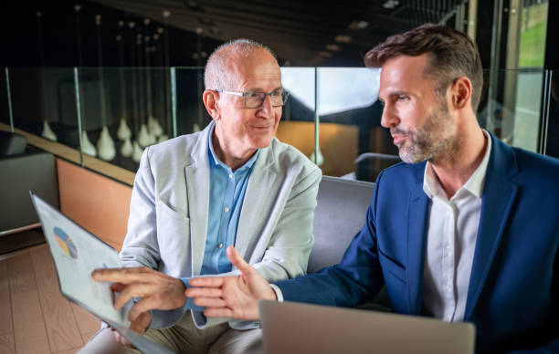 istock Younger and older businessman talking about documents during meeting in modern business lounge 1202965113