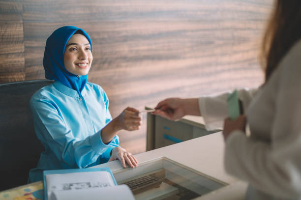 malaysia hospital contador asiático recepcionista femenino recibiendo el pago de la paciente femenina hiyab musulmana aceptando su tarjeta de crédito - healthcare and medicine receptionist paying credit card fotografías e imágenes de stock