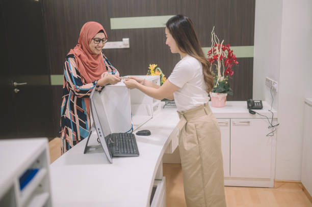 uma paciente asiática pagando sua conta hospitalar no balcão de registro com seu cartão de crédito entregue para o caixa feminino - dentist office built structure doctors office retail - fotografias e filmes do acervo