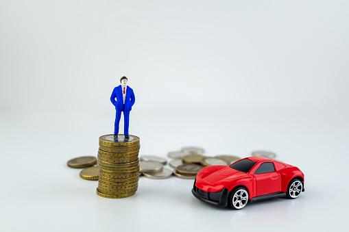 Miniature people: small businessman standing on stack of coins with a cute red car nearby, with some coins laying behind him on a white backround. He is rich. Money, Financial, Business Growth concept.
