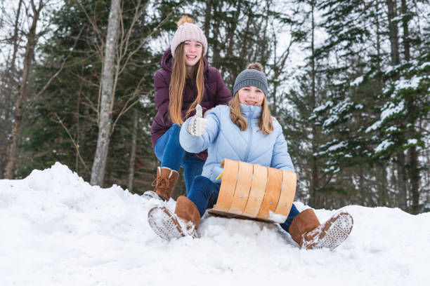 Two happy teen girls/friends sitting on a wood toboggan/sled while at the edge of a snow covered hill in a park and one girl holding her thumb up. stock photo