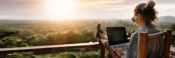 Photo of Young business woman working at the computer in cafe on the rock. Young girl downshifter working at a laptop at sunset or sunrise on the top of the mountain to the sea, working day.
