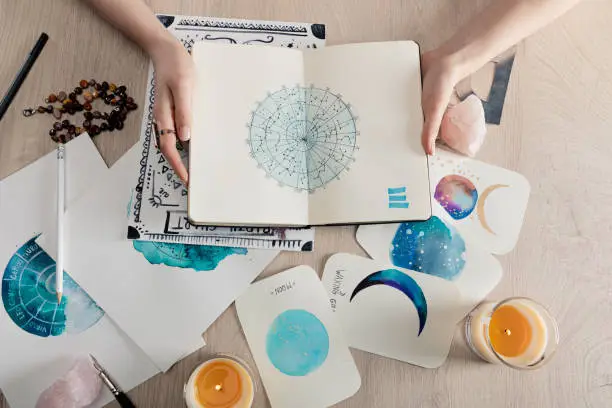 Photo of Top view of astrologer holding notebook with watercolor drawings and zodiac signs on cards on table