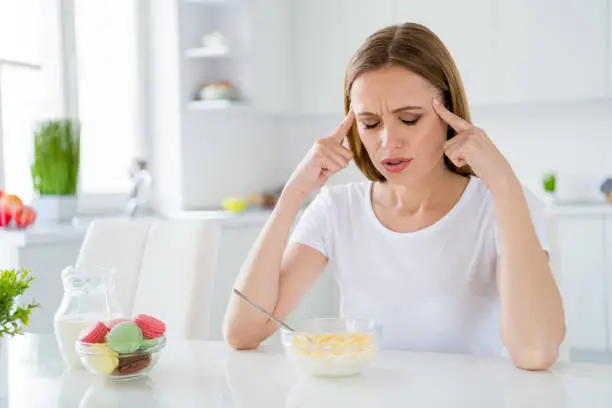 Photo of pretty housewife holding temples suffering terrible pain can't eat milk breakfast, cornflakes have hangover after night party sitting table white light kitchen indoors