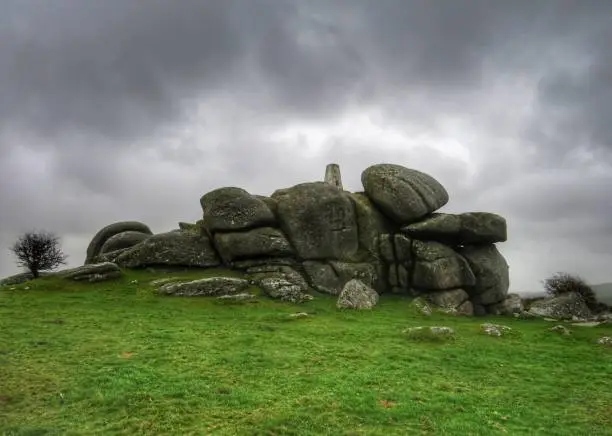 Dark, dramatic rainclouds hang heavy in the sky above Helman Tor, Cornwall, as the sun tries to burst through. The grey overcast sky is a stark contrast to the vibrant green grass.