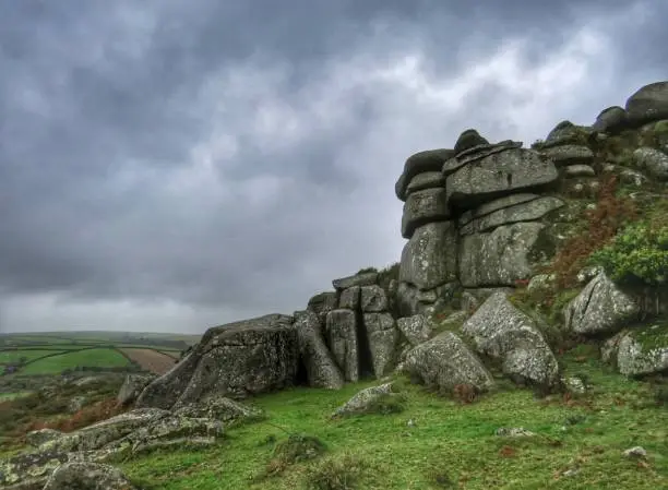 Dark, dramatic rainclouds hang heavy in the sky above Helman Tor, Cornwall, as the sun tries to burst through. The grey overcast sky is a stark contrast to the vibrant green grass.