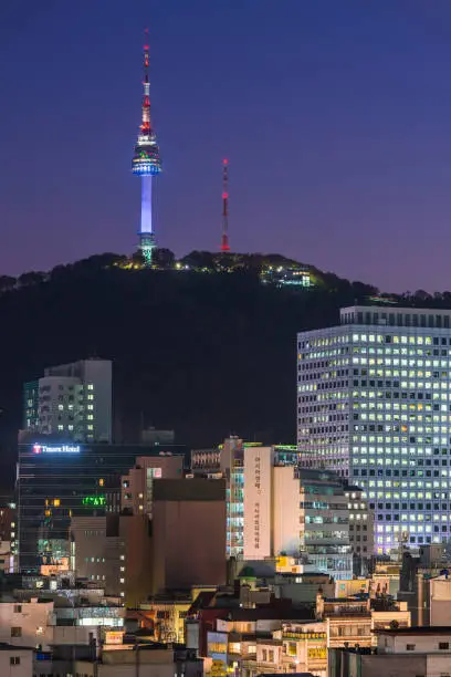 The iconic spire of Namsan Tower illuminated at sunset above the crowded high-rise cityscape of central Seoul, South Korea.