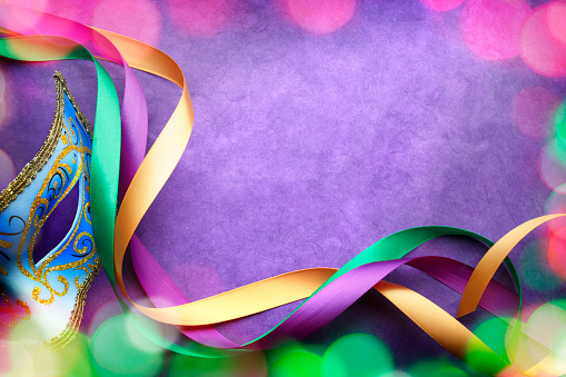 Colorful blurred lights surround a Mardi Gra mask and ribbons on a purple background that provides ample room for copy and text.