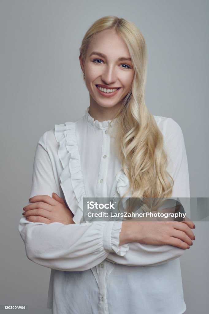 Young long-haired blond woman with her arms folded Young and beautiful long-haired blond woman in white blouse standing with her arms folded and looking at camera with a smirk. Front half-length portrait against grey background Application Form Stock Photo