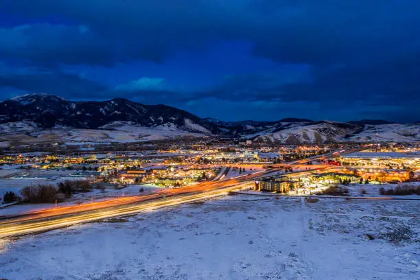 Aerial view of a snowy  Bozeman Montana at night. Retail stores and residential neighborhoods illuminate the landscape. Light trails on the freeway from cars.