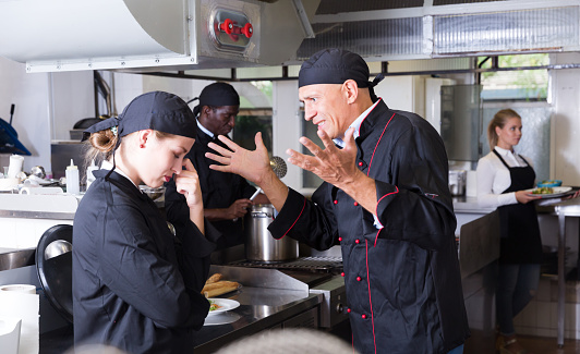 Enraged chef expressing dissatisfaction with work of frustrated girl in restaurant kitchen