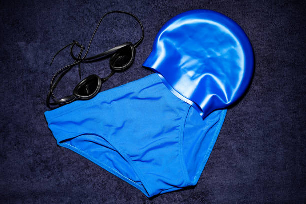 Blue men's swim gear on a navy blue towel. Swim briefs, swim cap, and swim goggles.