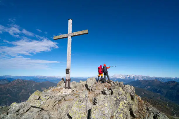 rear view of mountaineer couple standing together near summit cross on mountain peak on sunny but cold and windy day in autumn, man pointing with hiking pole, mountain range panorama in background