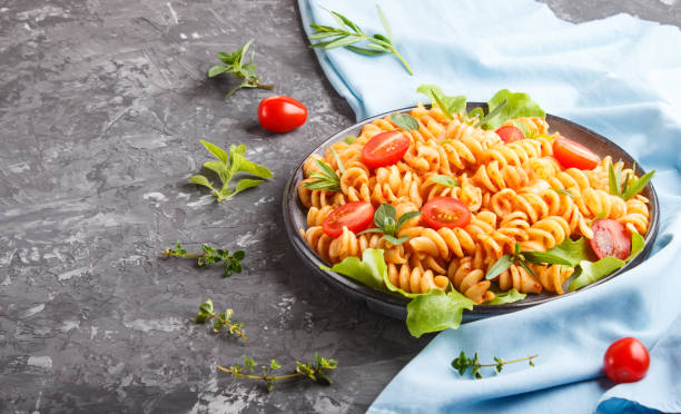 istock Fusilli pasta with tomato sauce, cherry tomatoes, lettuce and herbs on a black concrete background with blue textile. side view, copy space. 1203022717