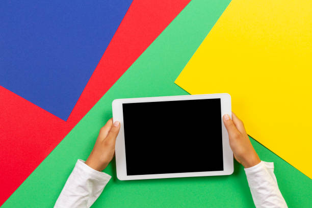 istock Kid hands holding white tablet computer on light green, blue, yellow and red background 1203024392