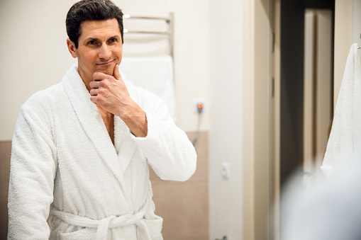 Joyful middle-aged man wearing a bath robe and looking at the camera while standing in hitel washroom
