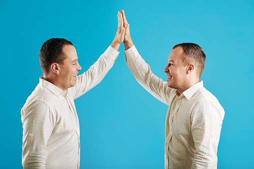 Young adult identical twin brothers wearing classic white shirts doing high five looking at each other smiling, blue background