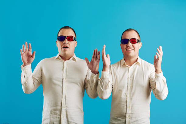Caucasian twin brothers wearing white shirts and anaglyph 3D glasses looking at camera, bright blue background