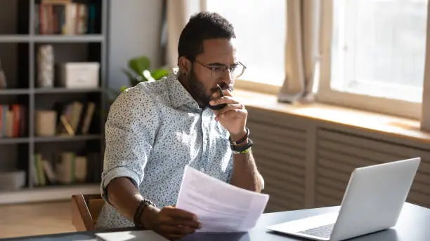 Photo of Focused african businessman holding documents looking at laptop doing research