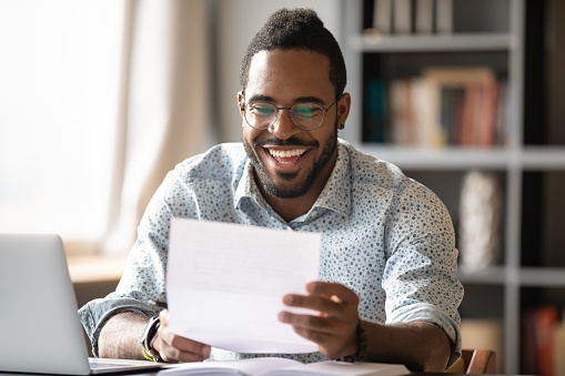 Cheerful happy young african american male student worker entrepreneur holding document bill read great news in letter receive loan approval scholarship salary payment concept sit at home office desk