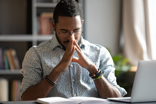 Young african american ethnic businessman praying asking for help concerned about problem making difficult decision concentrating mind meditating put hands in prayer sit at work desk in office