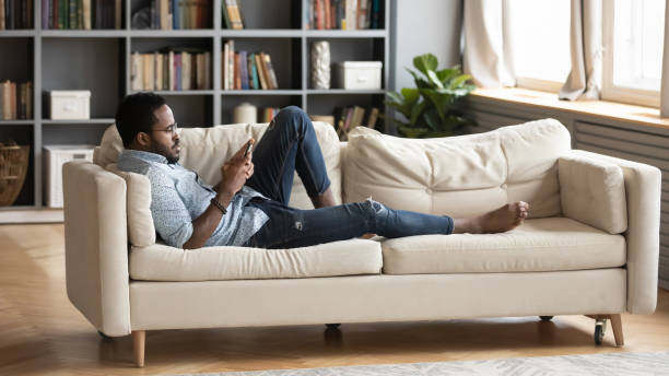 istock Serious relaxed millennial african man lying on couch using smartphone 1203044197