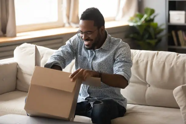 Photo of Smiling african man customer opening cardboard box parcel on sofa