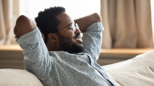 Happy african guy relaxing on couch eyes closed breathing air Happy serene african guy relax eyes closed breath fresh air chill on comfortable sofa, smiling lazy healthy ethnic young man resting on couch hold hands behind head lounge at home, side profile view relieved face stock pictures, royalty-free photos & images