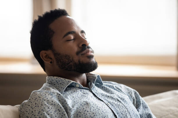 Tranquil young african man resting eyes closed breathing on couch Tranquil calm young african american man resting with eyes closed taking deep breath of fresh air enjoy comfort relaxing on couch feel peace mind and stress relief meditate at quiet home sit on sofa relieved face stock pictures, royalty-free photos & images