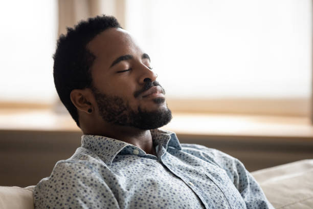 Tranquil young african man resting eyes closed breathing on couch Tranquil calm young african american man resting with eyes closed taking deep breath of fresh air enjoy comfort relaxing on couch feel peace mind and stress relief meditate at quiet home sit on sofa relief emotion stock pictures, royalty-free photos & images